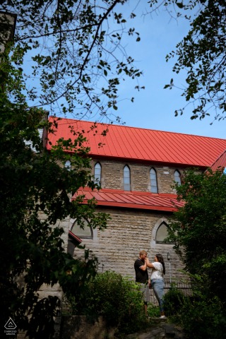At the Church of St. John the Evangelist in Montreal, QC, Canada, a man gently kisses his partner’s hand in an intimate pose. The couple is framed by trees, with the tall church building in the background.