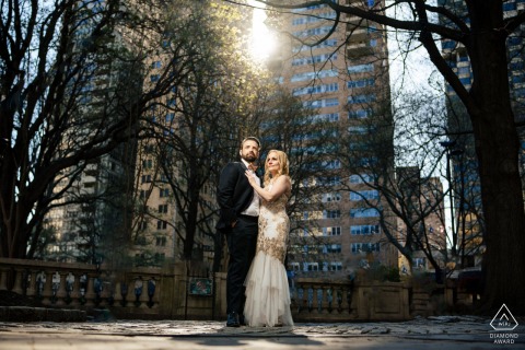 Radiant Love Shines In Rittenhouse Square Where City Skyline Meets Nature’s Beauty In Rittenhouse Square, Philadelphia, a couple stands together where nature meets the city skyline. The sun bursts brightly above and behind them, illuminating their love in this vibrant urban engagement portrait.