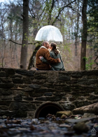 Romantic Couple Shares Kiss Under Umbrella On Stone Bridge At Wissahickon Valley At Wissahickon Valley Park in Philadelphia, a couple sits on stone bridge with their backs to the camera, sharing a kiss beneath a single umbrella. The river flows gently over rocks below, adding to the romantic scene.