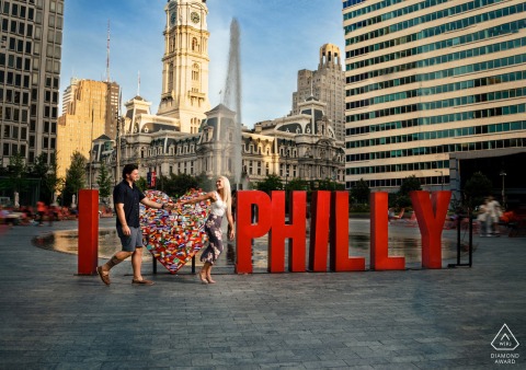 Love in Motion: Playful Couple Walks Briskly Through Philadelphia City Center At LOVE Park in Philadelphia, a couple walks hand in hand through the city center. She leads energetically, both moving briskly, capturing a dynamic and playful spirit in their urban engagement portrait.