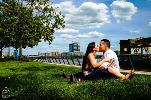 Relaxed Couple Shares Affectionate Smiles On Race Street Pier At Race Street Pier in Philadelphia, a couple sits with their legs tangled together on the grass, relaxed and connected. Their affectionate pose and easy smiles rest beneath the open sky in this romantic engagement portrait.