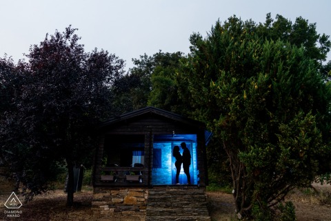 In Lugo, Galicia, Spain, a couple stands silhouetted and backlit by a blue spotlight, framed by trees. The dramatic lighting and natural surroundings create a striking pre-wedding engagement portrait.