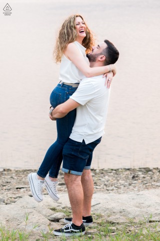 In Lugo, Galicia, Spain, a couple in jeans and white shirts enjoys a playful pre-wedding portrait. He lifts her up in a joyful hug, both smiling warmly in this lighthearted setting.