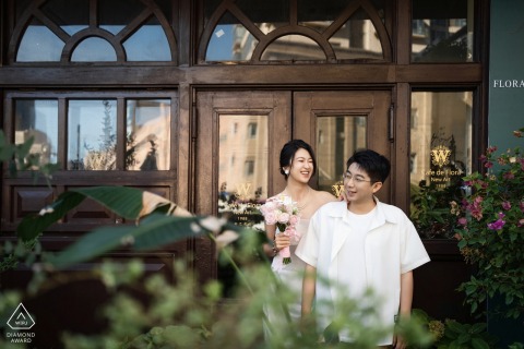 At the Bund in Shanghai, China, a couple shares laughter beside the door of a vintage flower shop, surrounded by lush greenery. Their joyful expressions capture a romantic session in an elegant urban setting.