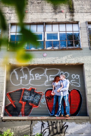 A loving couple shares a kiss on a San Francisco warehouse dock, with bright graffiti covering the old building's roll-up door.