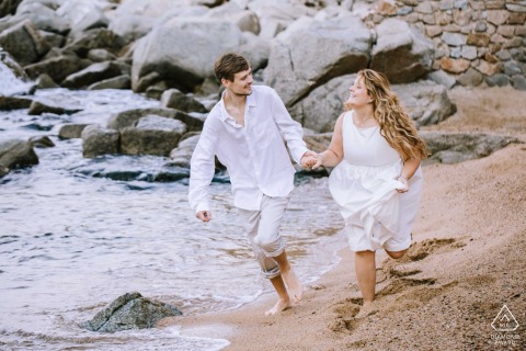 A couple runs joyfully on Platja d'es Codolar beach in Tossa de Mar, hands clasped, the Mediterranean Sea sparkling.