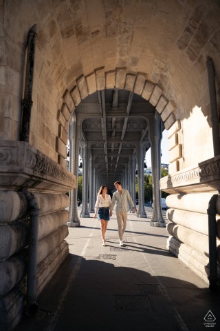 A couple stands hand in hand, looking at each other beneath the towering Bir-Hakeim bridge.