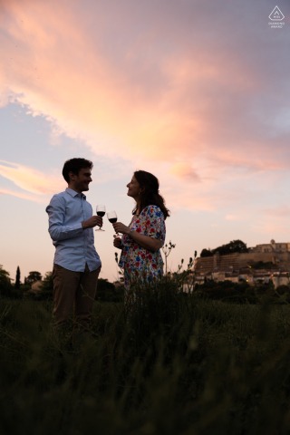 Drôme provençale, France engagement photography: Vertical frame of a couple toasting in Grignan. Romantic portrait with a soft pastel-colored sky in the background.