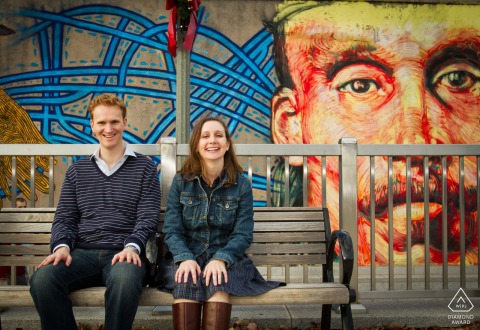 Decatur Square Mural Framing Confident Engagement: A Direct, Seated Portrait A deliberate and composed portrait of an engaged couple in Decatur Square, seated on a bench and framed by a vibrant street mural, presenting a confident, direct look to the camera.