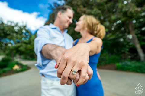 A shallow depth-of-field photograph showcasing an engagement ring in sharp detail. Using a wide lens, the couple is captured holding hands in the foreground, with the Denver Botanic Gardens' scenery blurred behind them.