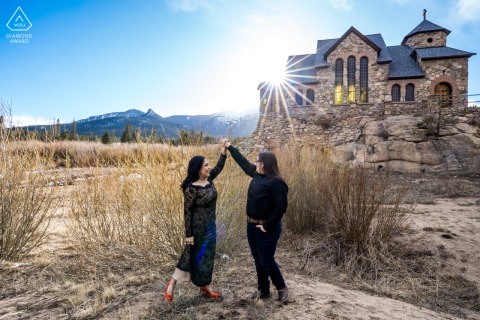 An engaged couple dancing near a castle-like structure in Allenspark, Colorado. The photograph features a dramatic sunburst directly behind them, set against a vast blue sky.