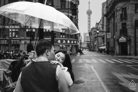 Engaged couple portrait in Shanghai, China on The Bund at night. The pair is sharing an intimate kiss under a transparent umbrella as soft rain falls on the iconic waterfront street. 
