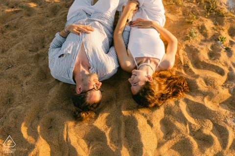An overhead portrait of an engaged couple lying on the sand at Les Sables d'Olonne, France. The camera captures them from above their heads, creating a unique upside-down perspective against the texture of the beach at sunset