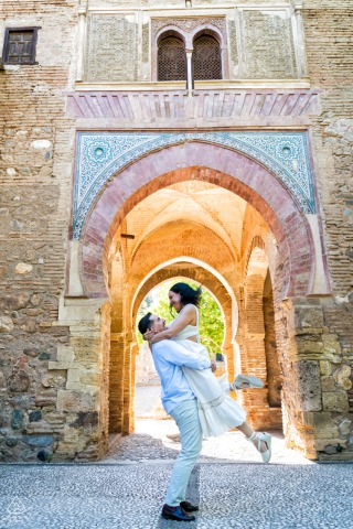 The groom-to-be joyfully picks up his fiancée off the ground at La Alhambra, Spain. Pastel arches of the old-world stone building glow behind them.