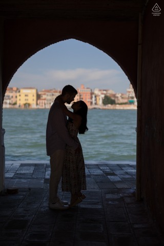 An engaged couple is silhouetted sharing a kiss on Giudecca Island, Venice. The figures are perfectly framed within an old-world stone arch, with bright blue water visible in the background. 