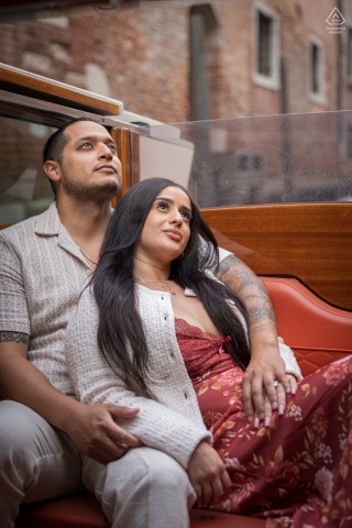 A newly engaged couple is seated on a Venice water taxi. She leans her back into him as they both look upward, sharing a peaceful journey through the Italian canals.
