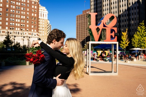 Philadelphia LOVE Embrace: Red Roses, Secure Hold, Forever Starts Now An engaged couple shares a close embrace in Philadelphia's LOVE Park. She holds a bouquet of red roses while wrapping her arms around his neck; he securely holds her waist.