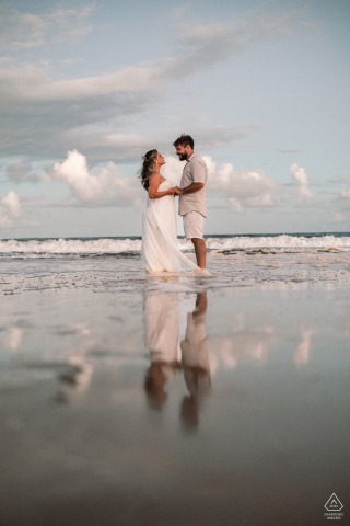A pre-wedding portrait from Praia de Ipioca, Alagoas. The couple's figures and the vast ocean are composed with their reflection in a water-mirror, emphasizing the tranquility of the coastal setting.