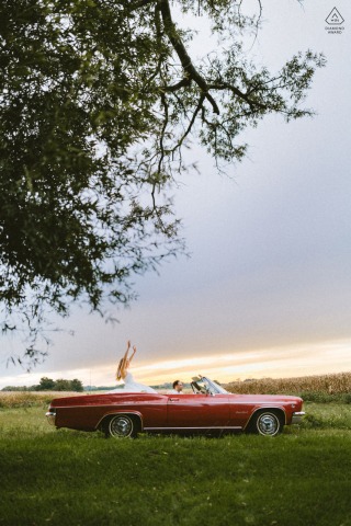 This lively sunset portrait in Gambrills, Maryland, shows a couple driving a red convertible vintage car; he drives while the lovely girl sits in the back, capturing a scene of fun times and shared adventure.