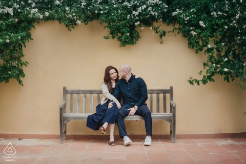 At Castello del Nero, Firenze, this Tuscany couple portrait captures the pair sitting close together on a wooden bench against a wall, ivy overhead framing them in a scene of quiet romance.