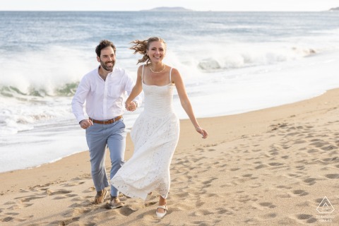 At Reid State Park in Georgetown, Maine, the couple runs joyfully on the sandy beach, smiling as ocean waves crash energetically behind them.