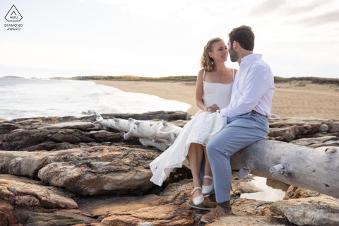 This lovely portrait at Reid State Park, Georgetown, Maine, captures the couple sitting close together on a large piece of driftwood by the water's edge on the beach, enjoying the coastal setting.
