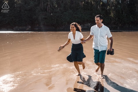 At Guarujá, Brasil, the couple dashes through shallow water, holding hands and their shoes, capturing a playful and spontaneous beach scene.