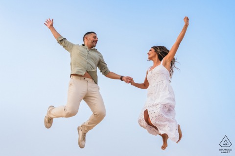 At Playa del Faro de Trafalgar, Cádiz, Spain, a pre-wedding photo captures the couple joyfully jumping together, both suspended mid-air against a minimalist, all-blue sky.