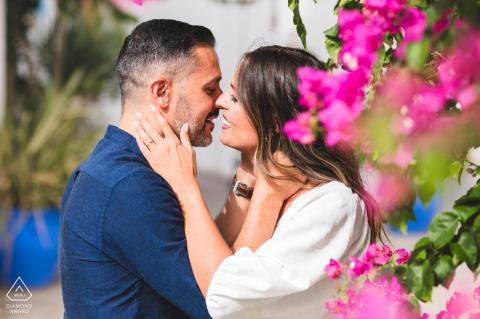 In Vejer de la Frontera, Cádiz, Spain, a pre-wedding portrait shows the couple face to face, about to kiss, with vibrant pink flowers filling the right foreground of the frame.