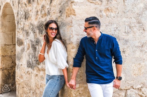 In Vejer de la Frontera, Cádiz, Spain, a pre-wedding photo shows the couple holding hands and smiling in sunglasses as they peek around a village alleyway corner, framed by Old World stone structures.