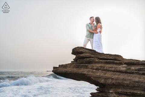 At Playa del Faro de Trafalgar, Cádiz, Spain, a pre-wedding portrait features the couple standing together on rocks overlooking the crashing waves of the sea.