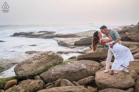 Playful Romantic Dip on Seaside Rocks in Playa del Faro de Trafalgar Couple Portrait Session At Playa del Faro de Trafalgar, Cádiz, Spain, a pre-wedding photo captures the couple sharing a playful and romantic dip on the rocks at the edge of the sea.