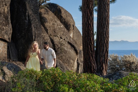 Walking the Scenic Trails by the Water in a South Lake Tahoe Engagement Portrait Session At South Lake Tahoe, CA, an engaged couple walks a scenic trail near boulders with stunning views of the water, celebrating their love amidst nature’s beauty.