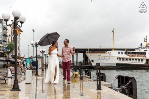 In Karaköy, Istanbul, a pre-wedding photo session shows her holding an umbrella as the couple walks together by the boat docks along the waterfront.