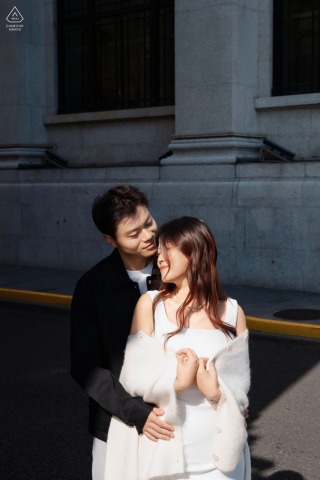 On the Bund in Shanghai, China, she is wrapped in his embrace, sunlight illuminating them while shadows fall on the Old World building behind, creating a romantic atmosphere.