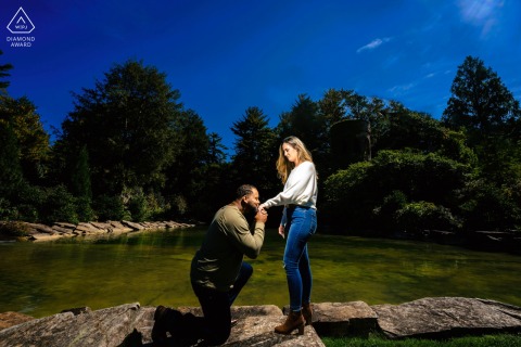 Engaged Couple Sharing a Tender Hand Kiss by a Pond in Philadelphia In Philadelphia, the couple shares a tender hand kiss beside a tranquil pond, surrounded by vibrant trees and a bright blue sky overhead.