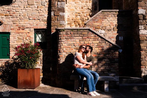Engaged Couple on Bench in Grado at Historic Building for Portrait Session In Grado, Gorizia, Italy, the couple sits together on a bench facing a beautiful, historic building, enjoying the charm and elegance of their surroundings.