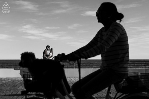 Engaged Couple Urban Portrait in Grado With Bicycle and Dog Silhouette In Grado, Gorizia, Italy, a couple portrait in black and white captures an urban street scene as the silhouette of a woman on a bicycle with her dog in the basket passes by in the foreground.