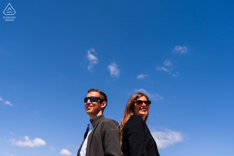 Engaged Couple Back to Back Under Blue Sky in Portopiccolo Portrait Session In Portopiccolo, Sistiana, Trieste, Italy, the couple stands back to back beneath a sunlit blue sky, captured in minimalist and symmetrical framing.