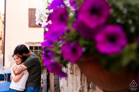 Engaged Couple Embraced Among Violet Flowers During Portrait Session in Grado In Grado, Gorizia, Italy, a couple portrait shows him hugging her from behind, with vibrant violet flowers framing the right foreground of the image.