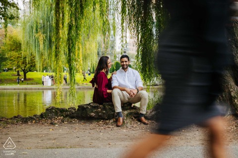 In Boston, MA, the couple sits close together by the water, enjoying the garden scenery as people stroll by in the background.