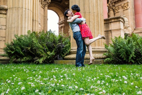 In San Francisco, CA, an engagement portrait from grass level captures the couple hugging and kissing, with an old-world stone building providing a romantic backdrop behind them.