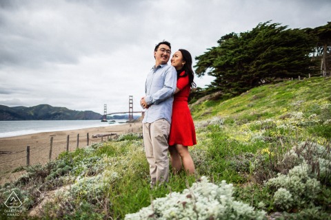 In San Francisco, CA, an engagement portrait shows her hugging him from behind as they stand among flowers, with the Golden Gate Bridge visible in the distance.
