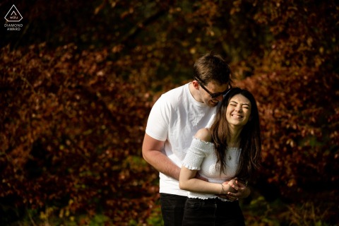 At Verulamium Park in St Albans, Hertfordshire, the bride- and groom-to-be laugh together in hard light with autumn trees in the background for their engagement portrait.