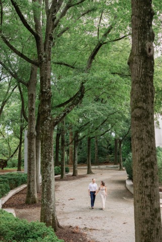 On Teddy Roosevelt Island in Washington, DC, a couple walks together between trees, captured in a tall vertical portrait with the wooded landscape framing the scene.