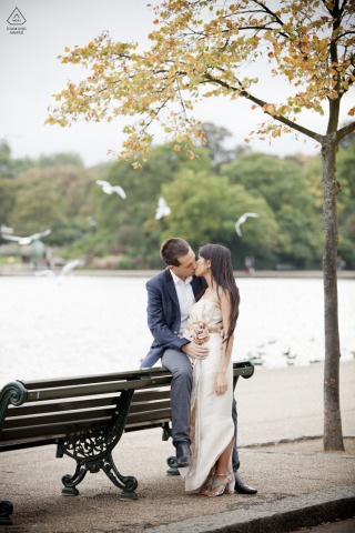 At Hyde Park in London, a couple kisses by the water with birds flying overhead, captured in a tall vertical portrait with trees framing the scene.
