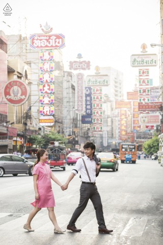 In Bangkok, Thailand, a couple holds hands while crossing a street, captured in a tall vertical portrait as they walk across a crosswalk.
