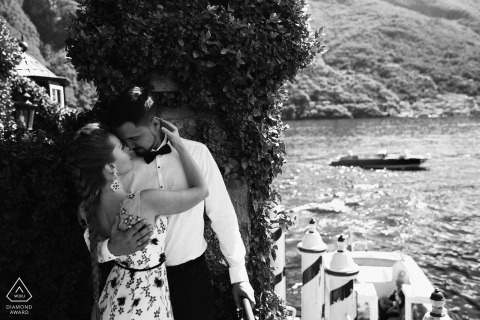 At Lake Como in Italy, a couple shares a quiet kiss beside the water in a black-and-white portrait captured from above the sea.