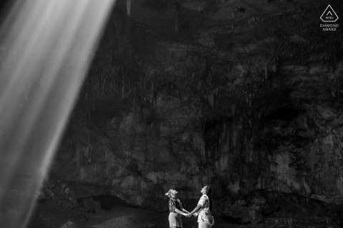 In Valladolid, a young engaged couple holds hands and faces each other inside a cave, looking up in a black-and-white minimalist portrait.