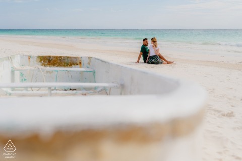 In Tulum, Mexico, a French couple sits together on the sand with blue water behind them, while a small fishing boat appears softly out of focus in the foreground for their portrait.